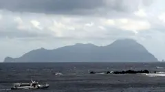 Akuseki Island, one of the main islands for tourists visiting Tokara, is seen in the horizon. A small ferry is in the sea, in the foreground ටෝකාරා නැරඹීමට පැමිණෙන සංචාරකයින් සඳහා වන ප්‍රධාන දූපතක් වන අකුසේකි දූපත ඈතින් දිස්වේ. මුහුදේ, ඉදිරිපසින් කුඩා තොටුපළක් ඇත.