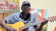 Umugabo wicaye mu nzu irimo irangi rijya kuba umweru, ahagatiye gitari arimo kuyicuranga, inyuma ye hari imitako ishushanyijeho inka n'intore. 