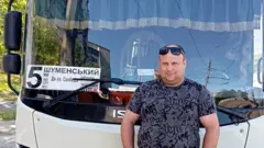 Mężczyzna stoi przed autobusem z numerem 5, ubrany w okulary przeciwsłoneczne i szarą koszulkę.