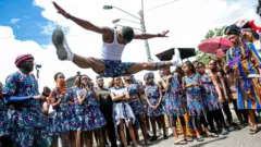 Une personne vêtue d'un gilet blanc et d'un short imprimé africain fait un grand écart en l'air tandis que des personnes vêtues du même tissu imprimé africain se tiennent autour et regardent.