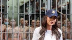 白いシャツ、帽子を着けたノーム氏が監獄の前に立っている。檻の後ろには、全身入れ墨のスキンヘッドの男性たちが多数いる