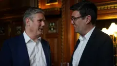 白いシャツに濃い色のジャケットを着た2人が向かい合って話している。木製のパネル張りの部屋に立っており、バーナム氏は手にグラスを持っている。
