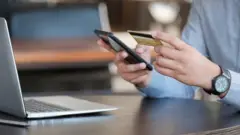 Pessoa com cartão de crédito e celular na mão em frente a laptop