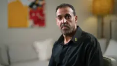 Portrait of Eli Sharabi in a black shirt with a yellow ribbon lapel pin, sitting in a room with largely neutral colours