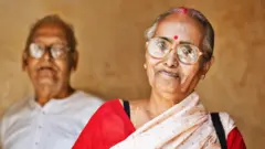 Um casal idoso. A mulher aparece em primeiro plano vestindo um sari vermelho e branco, e o homem, desfocado ao fundo, veste uma camisa branca.