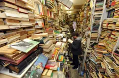 Jovem parado no meio de uma livraria antiga cercado por livros usados.