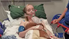 Megan está em uma cama de hospital com vários fios ligados a ela e uma máscara de oxigênio. Ela está de olhos fechados.