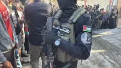 مقاتلون من حركة حماس يقفون استعداداً لتسليم الرهائن الإسرائيليين العشرين إلى فرق اللجنة الدولية للصليب الأحمر، ويظهر في الصورة مقاتل وجهه ملثم ويرتدي درعاً واقياً كُتب عليها "كتائب القسام"، وأعلى ذراعة شارة عليها وجه ملثم وكتب عليها "وحدة الظل"، بينما يحمل سلاحاً نارياً.
