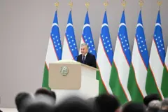 Shavkat Mirziyoev minbarda nutq so'zlamoqda. orqasida O'zbekiston bayroqlari.