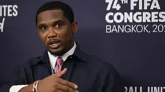 Samuel Eto'o fait un geste du bras droit alors qu'il est vêtu d'une chemise et d'une cravate et qu'il se tient devant un panneau d'affichage indiquant 74e Congrès de la Fifa à Bangkok.