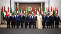 Desetine muškaraca u crnim i plavim odelima i jedan u beloj dugoj tunici pozira pred kamerom, iza njih su zastasve arapskih država koje predstavljaju