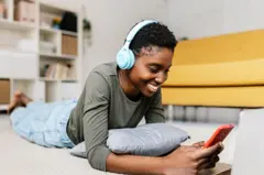 Femme noire utilisant un smartphone et des écouteurs alors qu'elle est allongée sur le sol de sa maison. 