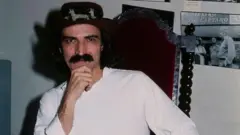 Belchior sentado em cadeira com olhar confiante para a câmera