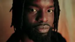 Un gros plan de Meji Alabi, barbe et dreads, regardant la caméra