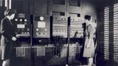 Fotografia em preto e branco mostra duas mulheres diante de um grande computador de primeira geração, cercado por painéis de controle, cabos e fileiras de componentes eletrônicos, em uma sala de processamento de dados.