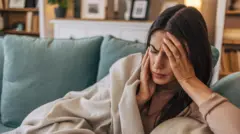 زنی با پتو روی مبل نشسته با دستش سرش را گرفته در حالیکه به نظر می‌آید سردرد دارد