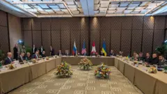 Negocjatorzy siedzą przy stołach w wyłożonym panelami pomieszczeniu, z flagami Rosji, USA, Szwajcarii i Ukrainy w tle.