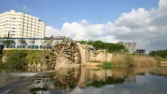صورة تظهر ناعورة تُستخدم لجر مياه النهر، ومن خلفها تظهر مبانٍ حديثة.
