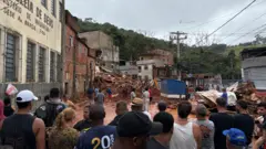 曇り空の下、緑の木々と斜面を背に、二階建てや三階建ての建物が並び、そのひとつが大量の赤い土砂とともに崩れている。重機が入って捜索活動が続く様子を、手前に集まった大勢が見ている