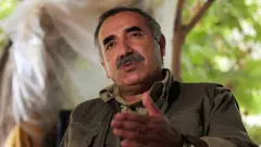 Koyu yeşil giysili bıyıklı bir adam konuşuyor. 