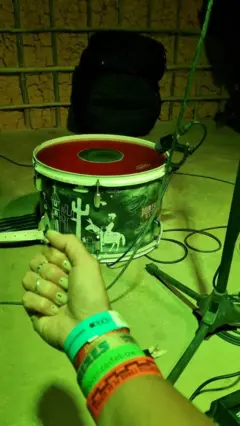 Braço de Priscila com diversas pulseirinhas de festivais de forró, com uma zabumba e uma parede de taipa ao fundo