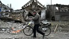Мужчина с велосипедом идет мимо разрушенных зданий в поселке на границе Сумской области 