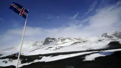 Uma bandeira islandesa tremula no alto de um mastro na encosta de uma montanha nevada.