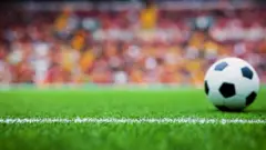 Yeşil sahada futbol topunu görüyoruz, dolu tribünler odak dışında. 