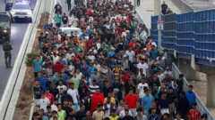 Um grande grupo de migrantes, principalmente homens, sendo escoltado pela polícia ao longo de uma passarela.