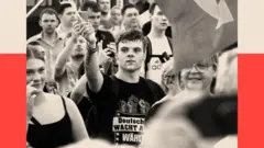 Толпа людей с флагами, в центре кадра молодой мужчина в футболке с изображением сжатых кулаков, он тоже держит флаг.