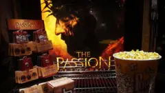 Uma bancada de cinema exibe baldes de pipoca e produtos promocionais diante de um pôster iluminado do filme A Paixão de Cristo. A imagem tem tons quentes e escuros, criando uma atmosfera dramática que remete ao clima intenso e religioso do longa-metragem.