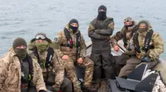 Six men sat on a boat on the ocean wearing army uniform and balaclavas look at the camera