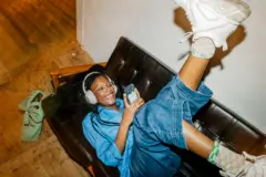 Mulher com fone de ouvidos deitada em um sofá com as pernas para cima, claramente se divertindo enquanto olha seu telefone celular.