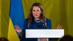 Христя Фріланд виступає за трибуною на тлі українського прапора