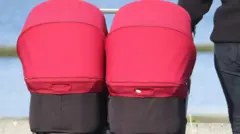 Um carrinho de bebê para gêmeos, de cor vermelha, sendo empurrado por um dos pais