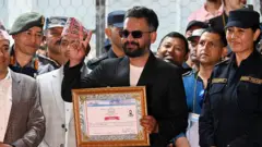 暗い色のサングラスをかけ、あごひげを生やした黒っぽいジャケット姿の男性が、右手でピースサインを見せ、左手に額入りの証明書らしきもの持って胸の前で見せている。横や後ろに多くの人が立っている。みんなほほ笑んでいる