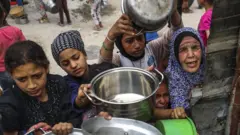 Palestinos, lutando contra a fome devido ao bloqueio israelense, esperam na fila com panelas vazias nas mãos para receber refeições distribuídas por organizações humanitárias em Gaza, em 17 de maio de 2025