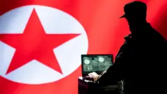 ከሰሜን ኮሪያ ባንዲራ ፊት ለፊት ተቀምጦ ላፕቶፕ ላይ እየሰራ ያለ ኮፍያ ያደረገ ሰው