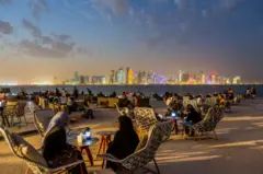 Pessoas sentadas em mesas de restaurantes, com prédios iluminados atrás, ao anoitecer