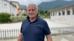O prefeito Giacomo de Luca, um homem com uma camisa polo azul marinho, está em frente aos prédios