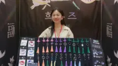 Choi Nyun-hee sorri para câmera exibindo um painel com artesanatos feitos por ela
