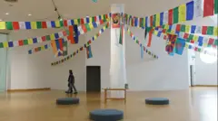 A large pillar stands in the middle of a gallery wrapped in white cloth, with several strings of colourful bunting carrying the flags of various countries and Tibetan art tied to the top of the pillar where a red and yellow flag has been pinned. There is a small wooden table at the base of the pillar as well as black circular cushions placed around it. Two people dressed in jeans and dark T-shirts are walking across the gallery