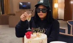 Brenda Biya porte un toast, vétue d'un sweat noir et portant une casquette vert olive et des lunettes noires. Elle est assise dans la chambre d'un hotel. 