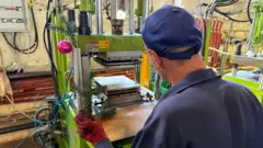 Homem de costas usa uma máquina para produzir decorações natalinas, com o ramo de uma árvore de Natal na mão e uma bola rosa pendurada na máquina