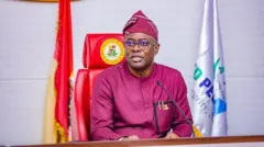 Gomina Seyi Makinde jokoo lori aga gomina rẹ to ni asia ijọba Oyo lapa ọtun ati asia ẹgbẹ oṣelu APM lapa osi. Ẹrọ gbohungbohun wa niwaju rẹ, bẹẹ lo de igo soju, lasiko to n sọrọ lọwọ