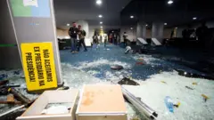 Detalhe do Congresso Nacional com vidros quebrados no chão e um cartaz: "Não aceitamos ex presidiários na presidência!".