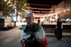 مؤيد للحكومة الإيرانية أسمر البشرة لديه لحية خفيفة بيضاء اللون، يرتدي العلم الإيراني يرفع يديه داعياً.