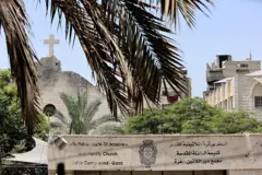 صليب على كنيسة متضررة وأمامها مكتوب على لوحة أسمنتية "البطريركية اللاتينية كنيسة السائلة المقدسة مجمع دير اللاتين -غزة"، باللغتين العربية والإنجليزية. وهناك عدد من الأشجار. الصورة التُقطت نهاراً. 