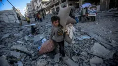 Израил Газадагы Абу Хасира көчөсүнө чабуул койгондон кийин палестиналык жаш балдар жарактуу буюмдарын терип жатышат. Арт жакта бир нече имарат толугу менен кыйраганын көрүүгө болот. 2025-жылдын 30-сентябры