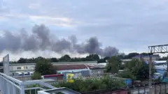 Промислова зона з кількома будівлями та густими клубами темного диму, що підіймаються над деревами на горизонті.